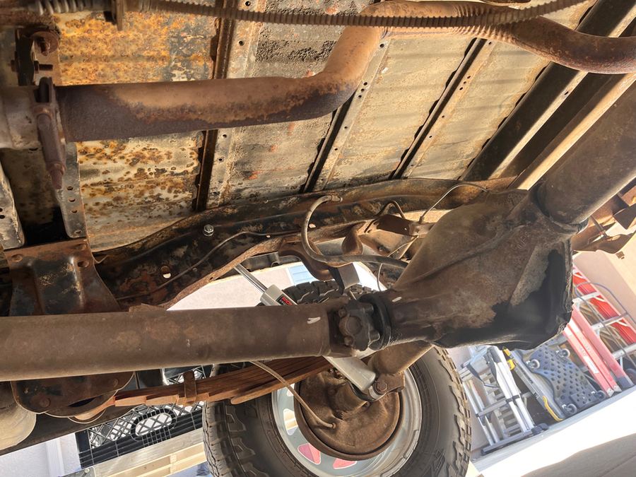 Underside of a pickup showing a rusted floor pan, driveshaft and rear axle