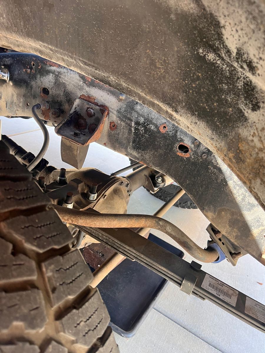 Rusted frame rail and spring hanger bracket behind the front wheel of a pickup