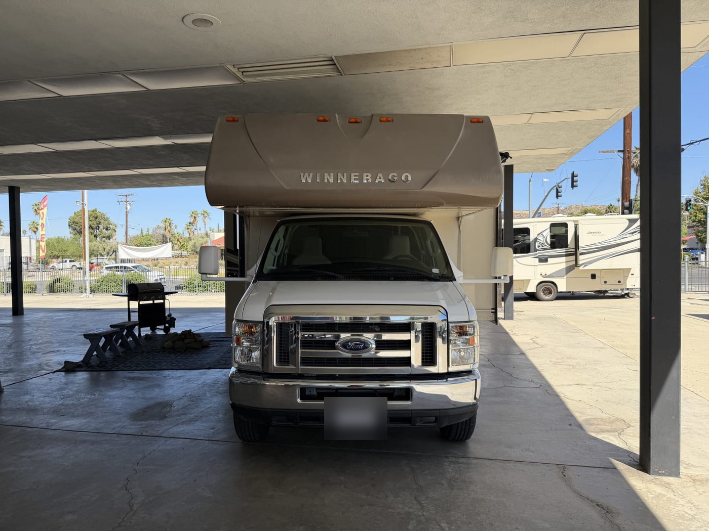Front of a Winnebago Class C motorhome on a Ford chassis under a carport, plate blurred