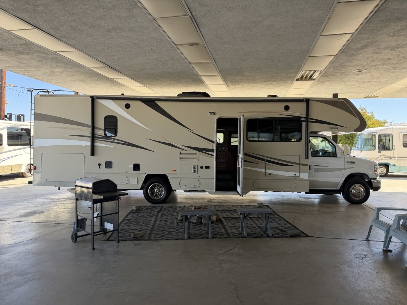 Door side of a Winnebago Class C motorhome under a carport, entry door open and a grill out front