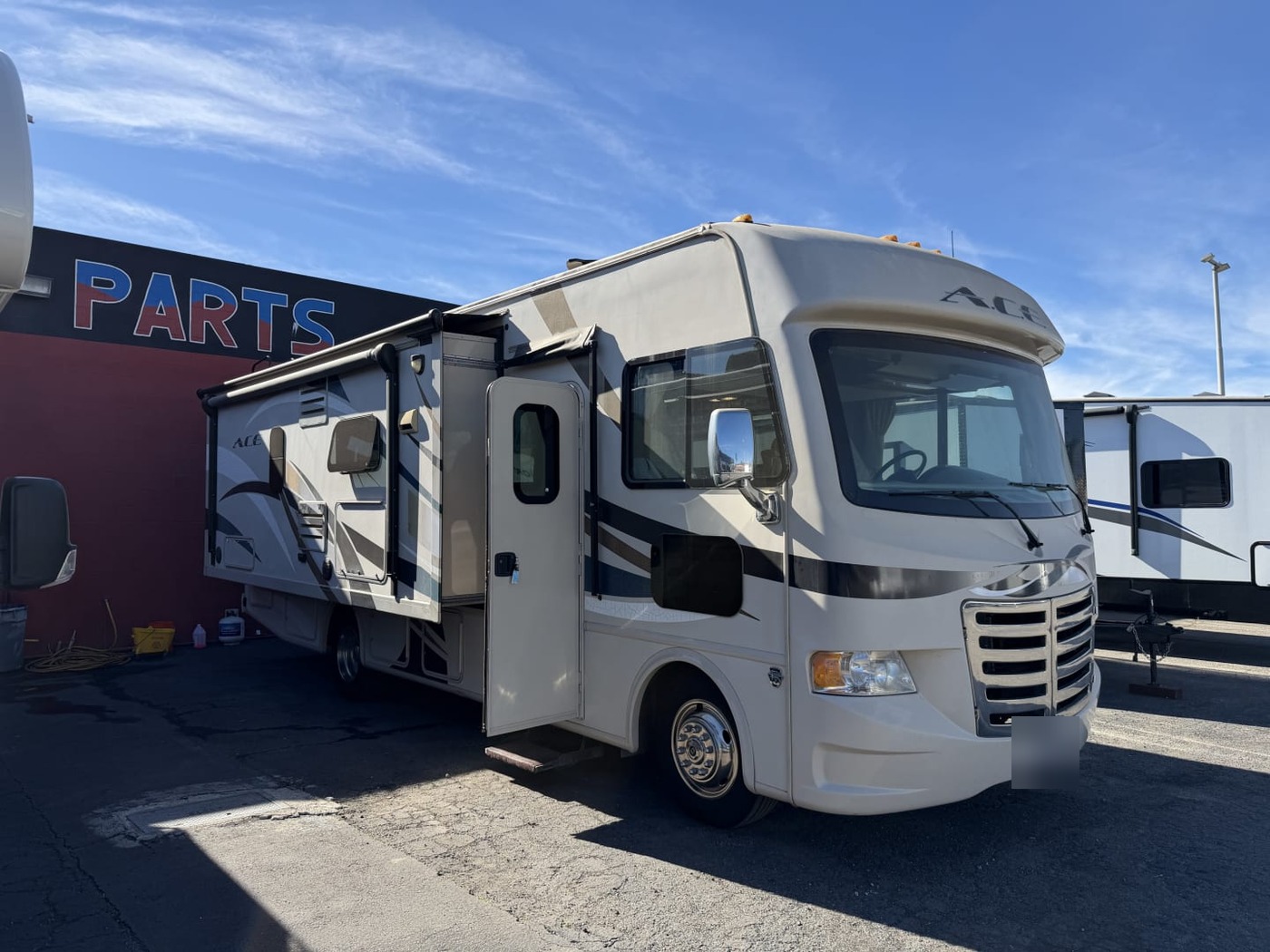 Tan Thor A.C.E. Class A motorhome parked on a lot with its entry door open, plate blurred