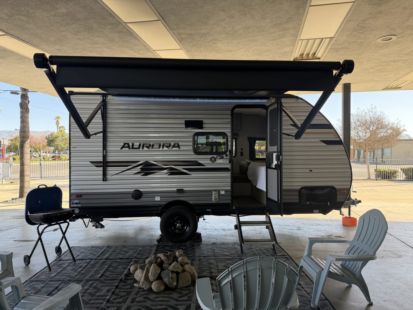 Forest River Aurora travel trailer under a carport with its awning extended and camp chairs in front